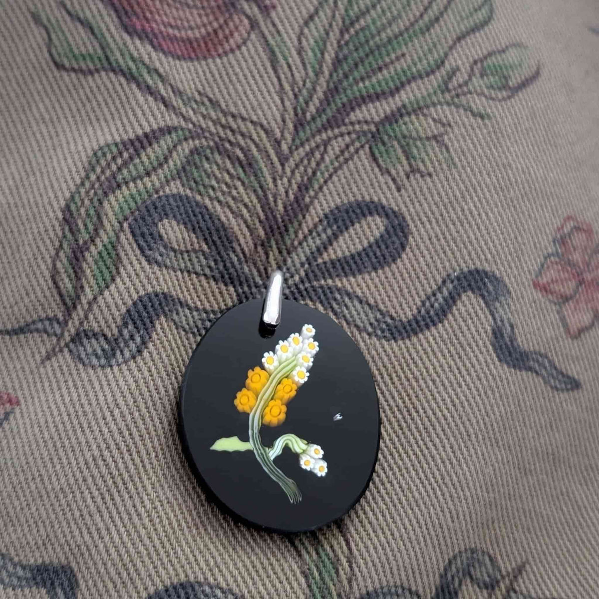 A round black pendant featuring yellow and white flower design, resting on a patterned fabric background.