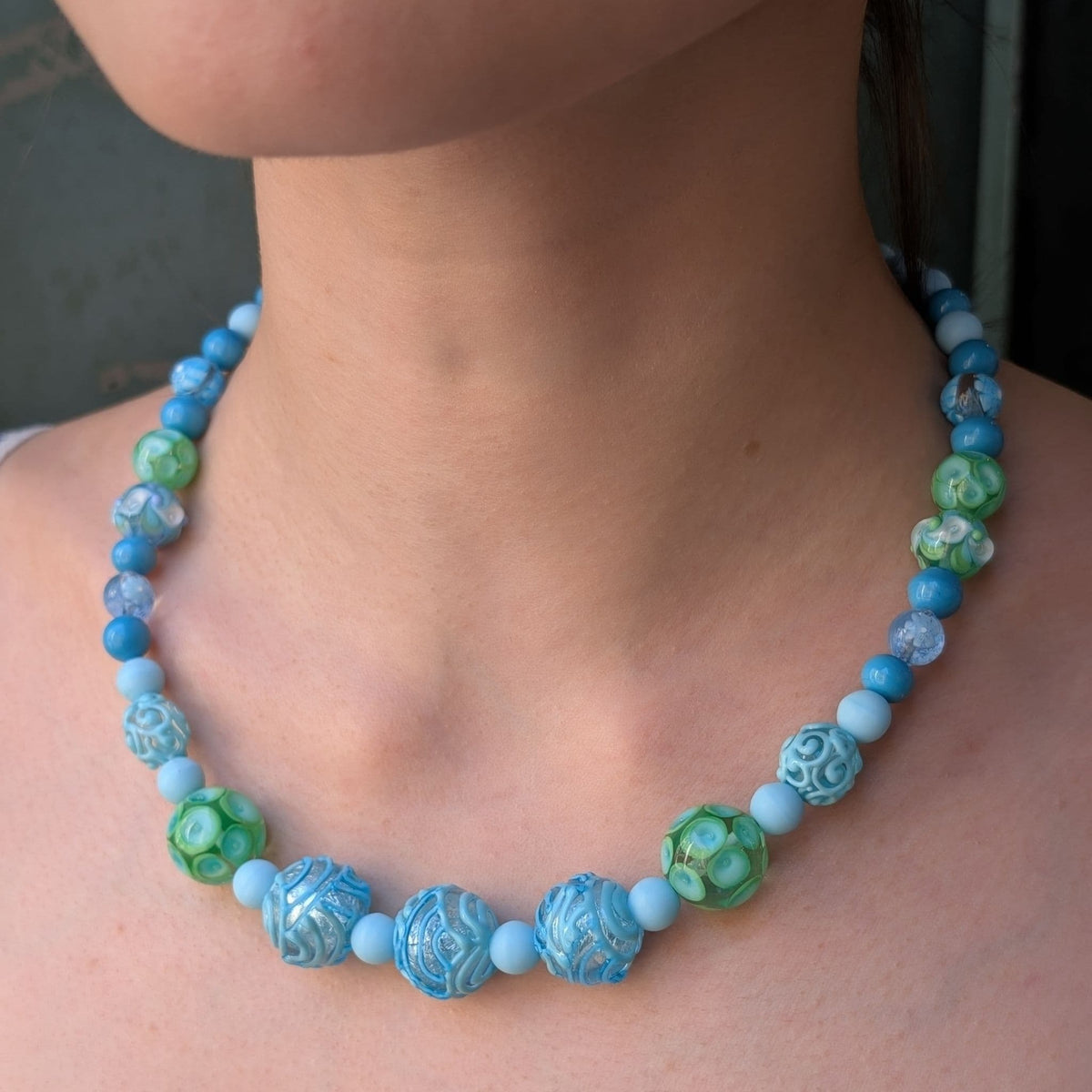 A close-up of a person's neck and upper chest wearing a beaded necklace with light blue, turquoise, and green marbled beads.